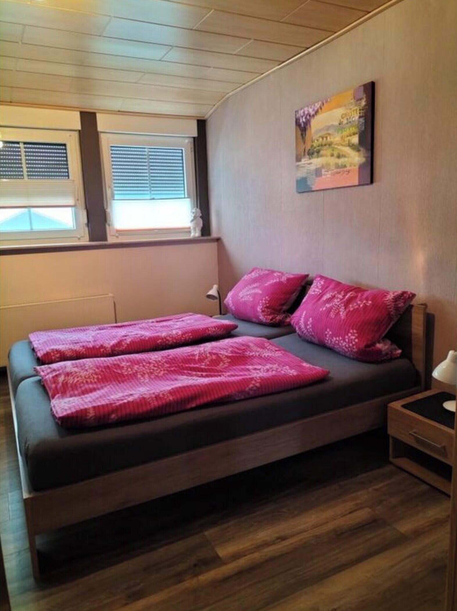 A bedroom with a wooden bed, two pink pillows, a nightstand, and a window with blinds.
