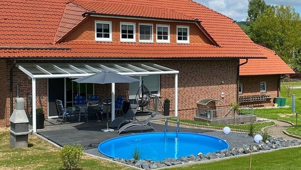 Ein zweigeschossiges Haus mit rotem Ziegeldach, einem Swimmingpool und einer überdachten Terrasse mit Gartenmöbeln.