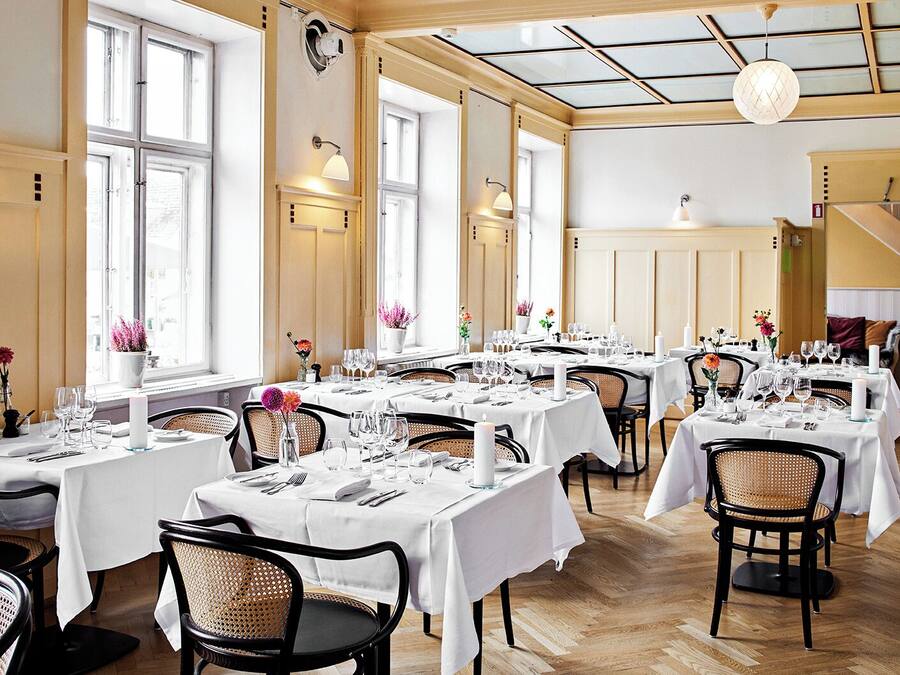 A restaurant with multiple tables set for dining, featuring white tablecloths, wicker chairs, and floral centerpieces.