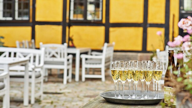 A tray of champagne glasses on a wooden table in an outdoor seating area with white chairs and a yellow and black building in the background.