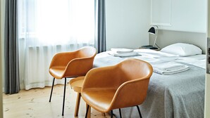 A hotel room with a bed, two chairs, a bedside table with a lamp, and a window with curtains.