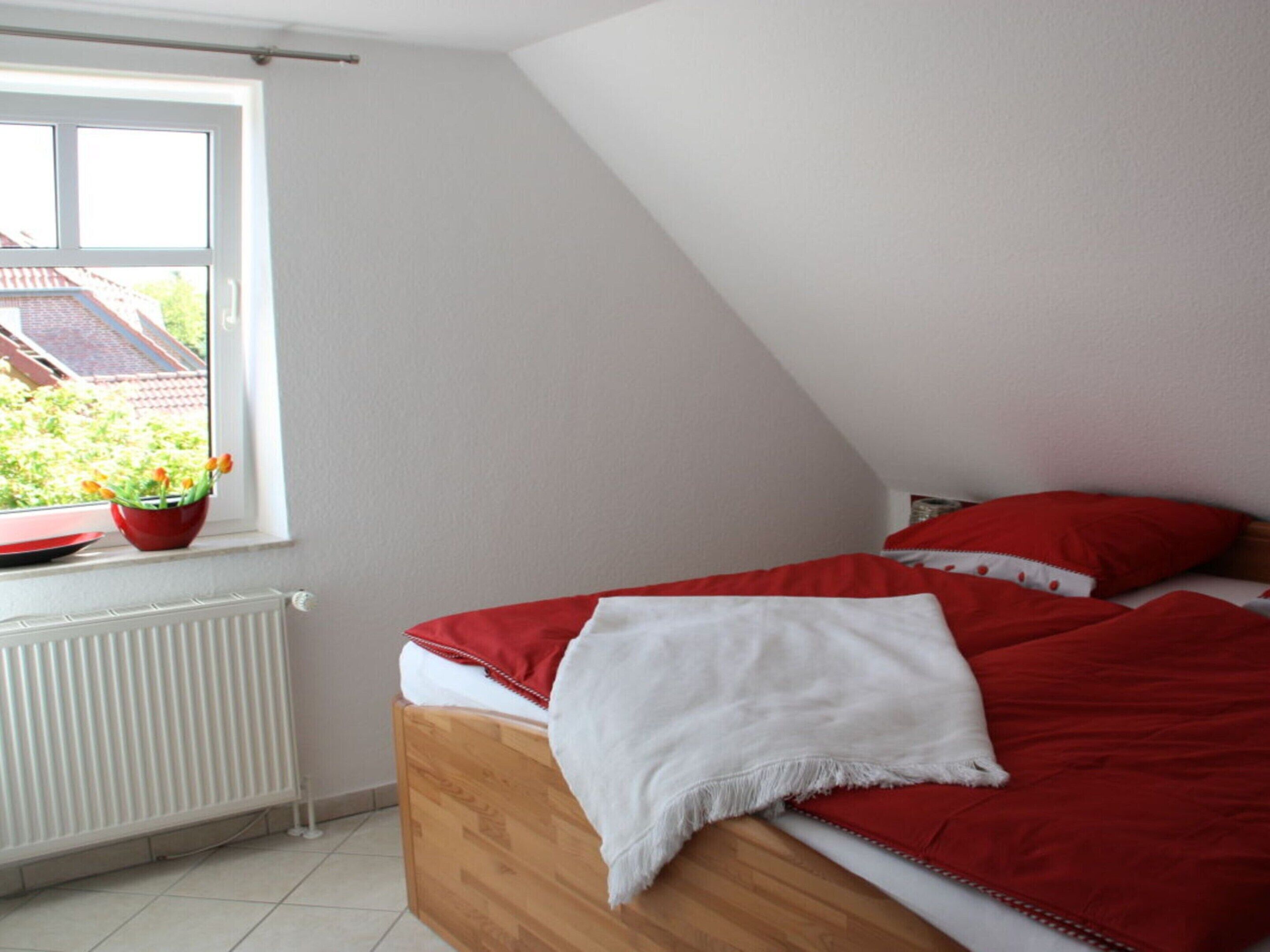 A bedroom with a sloped ceiling, a wooden bed with red and white bedding, a window with a view of a house and greenery, and a radiator under the window.