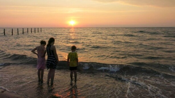 Atardecer sobre el océano con tres personas de pie en aguas poco profundas.