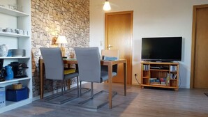 Una sala de estar con una pared de piedra, una mesa de comedor con sillas, un televisor sobre un mueble de madera y estanterías con libros y utensilios de cocina.