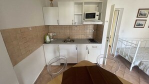 A compact kitchen with white cabinets, a microwave, a sink, and a small countertop. There are two clear plastic chairs and a bed with a metal frame in the adjacent room. - Apartment G&G Red with swimming pool (Poreč)