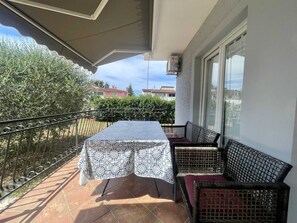 A balcony with a table and two wicker chairs, overlooking a garden and neighboring buildings.