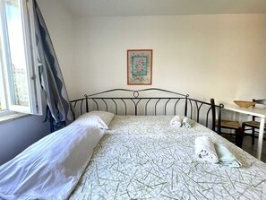 A neatly made bed with a patterned bedspread, a pillow, and rolled towels on a metal headboard. A framed picture hangs on the wall above the bed.