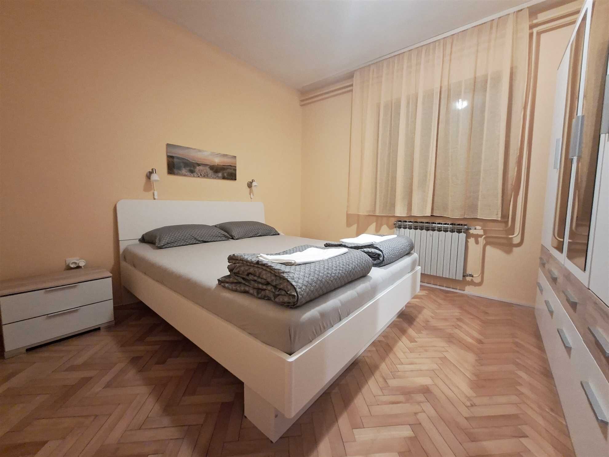 A bedroom with a bed, a nightstand, a radiator, and a window with curtains.