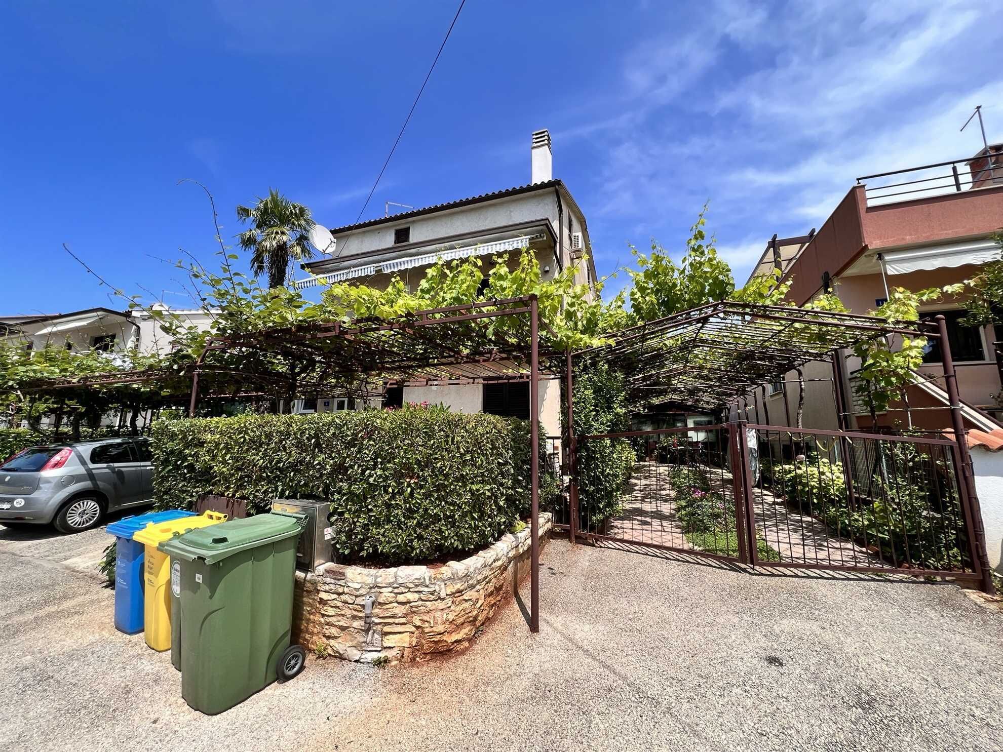 Il s’agit d’un quartier résidentiel où se trouve une maison dotée d’un mur en pierre et d’une pergola couverte de plantes grimpantes. On peut voir trois poubelles devant la maison.