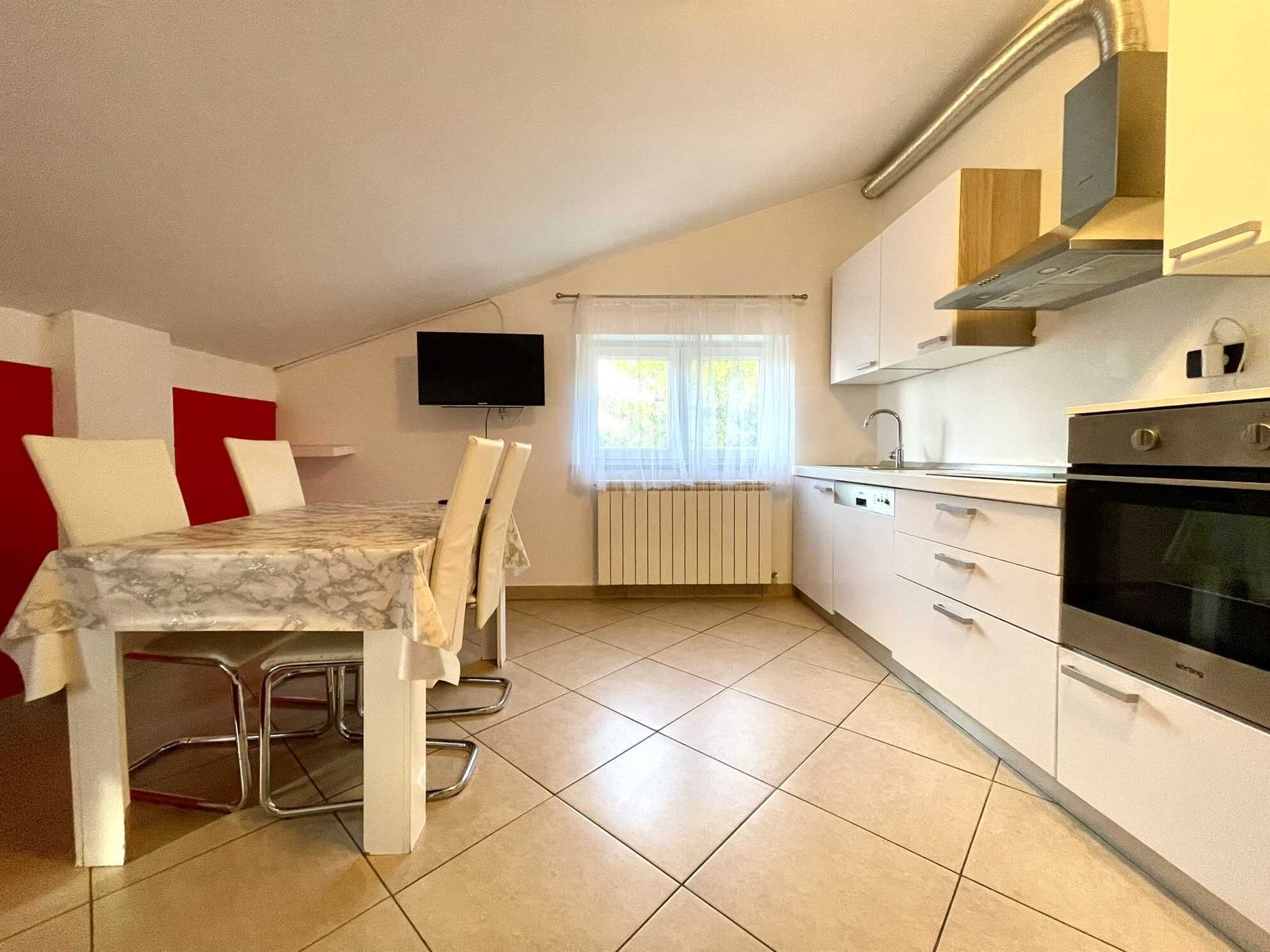 A compact kitchen with a dining area, a TV mounted on the wall, and a window with sheer curtains.