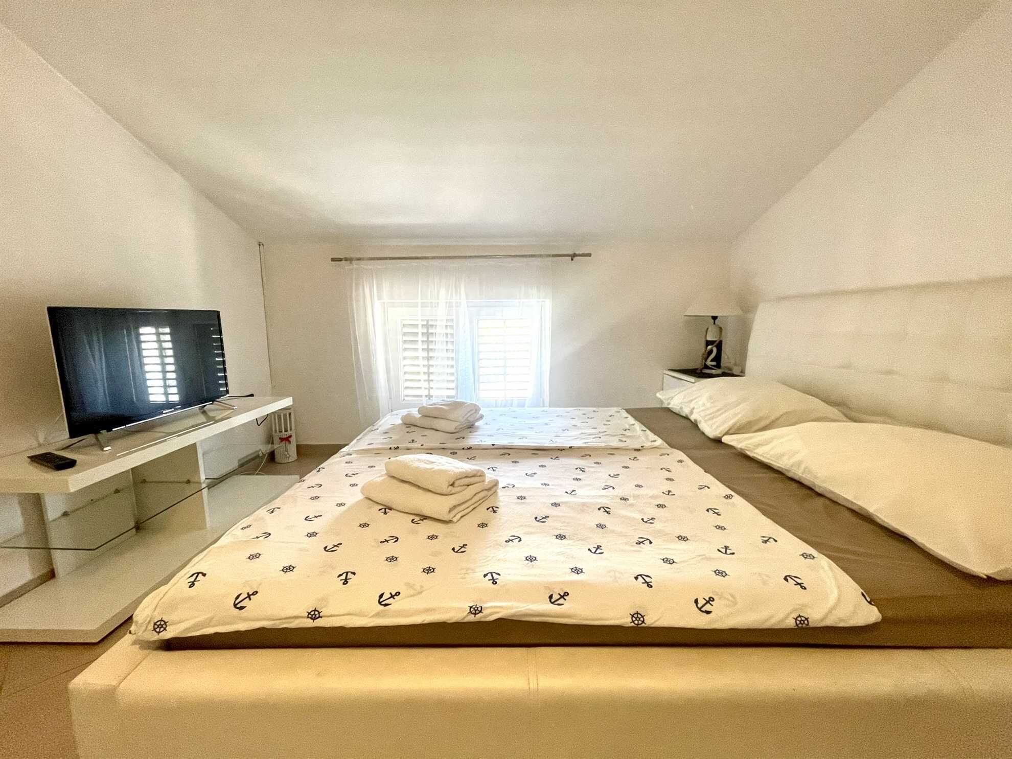 A hotel room with a TV on a stand, two beds with matching bedding, a bedside table with a lamp, and a window with blinds.