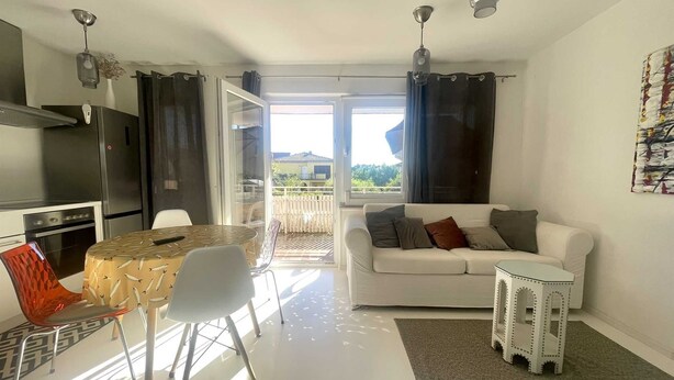 A modern living room with a white sofa, a dining table with chairs, a kitchen area with a refrigerator, and a sliding glass door leading to a balcony. - Apartment Mariana 1 close to the beach (Umag)