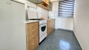 Een kleine keuken met een ingebouwde oven, een fornuis en een magnetron. Er zijn houten kastjes en een raam met zonwering.