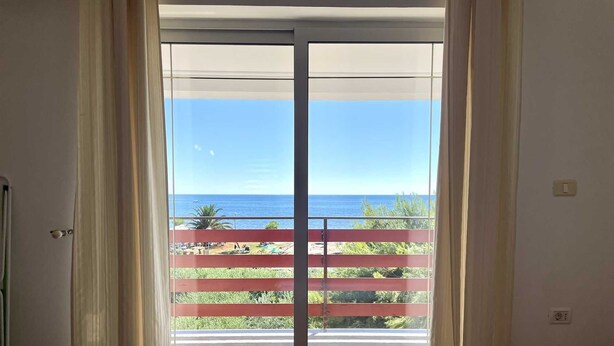 Quarto com porta de vidro deslizante que dá para uma varanda com vista para o mar e guarda-corpo vermelho.