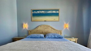 Quarto com cabeceira de madeira, colcha estampada, duas almofadas iguais e um quadro de uma praia acima da cama.