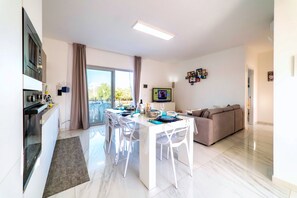 Uma cozinha moderna com micro-ondas embutido, um espaço de refeições com mesa e cadeiras brancas, e uma sala de estar com sofá.