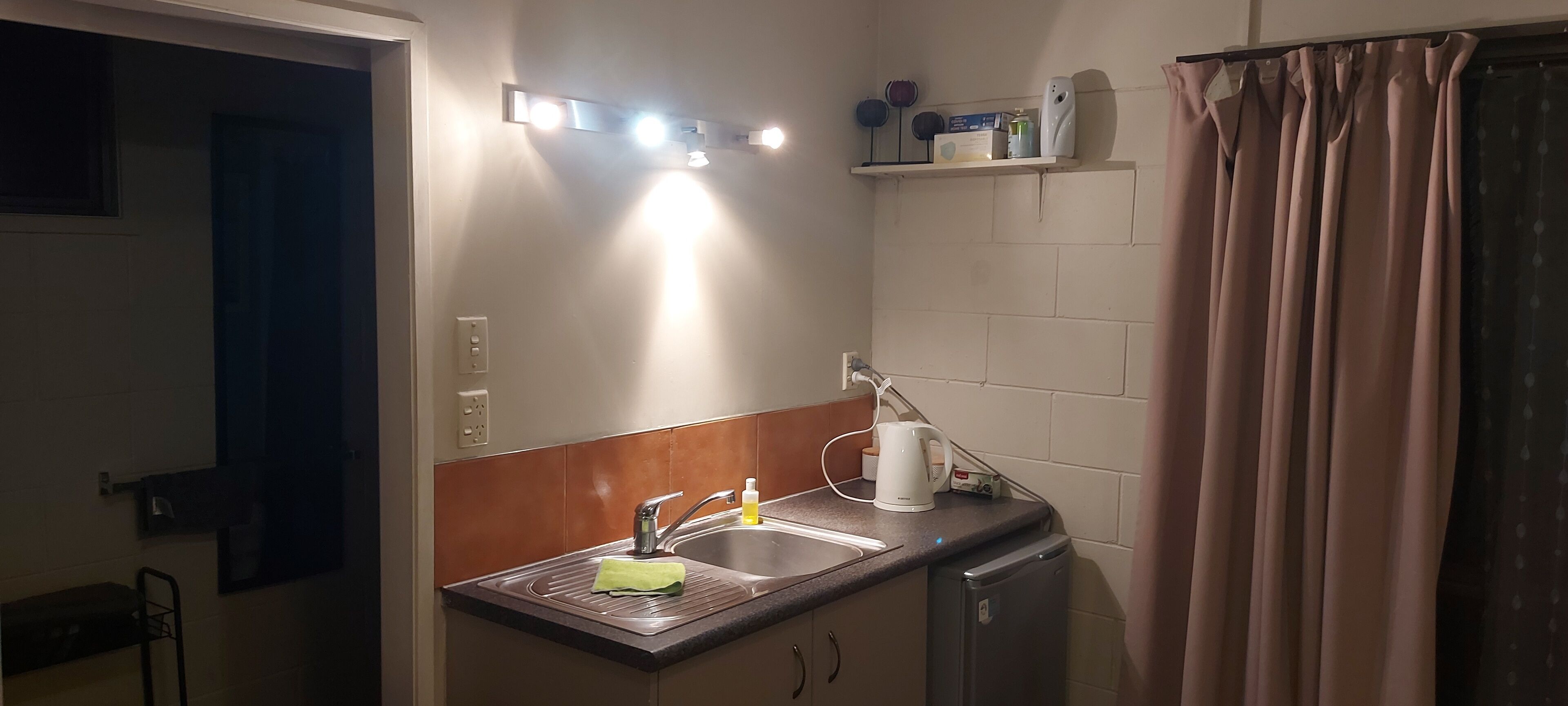 A compact kitchen with a sink, a kettle, and a small refrigerator. There is a shelf above the sink with a few items on it.