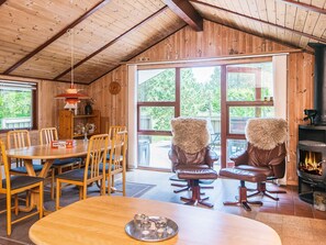 A cozy wooden cabin interior with a dining area, two leather chairs with fur throw pillows, a fireplace, and large windows offering a view of the outdoors.