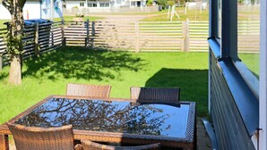 A patio with wicker furniture and a glass table, overlooking a grassy area with a wooden fence and houses in the background. - 4 Star Holiday Home in Skummeslovsstrand (Skummesloevsstrand)