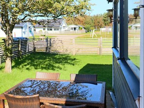 A patio with wicker furniture and a glass table, overlooking a grassy area with a wooden fence and houses in the background. - 4 Star Holiday Home in Skummeslovsstrand (Skummesloevsstrand)