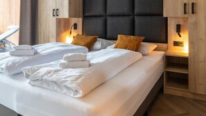 Un lit bien fait, avec du linge de lit et des serviettes blanches, une tête de lit rembourrée de couleur foncée et des meubles en bois équipés d’un éclairage mural.