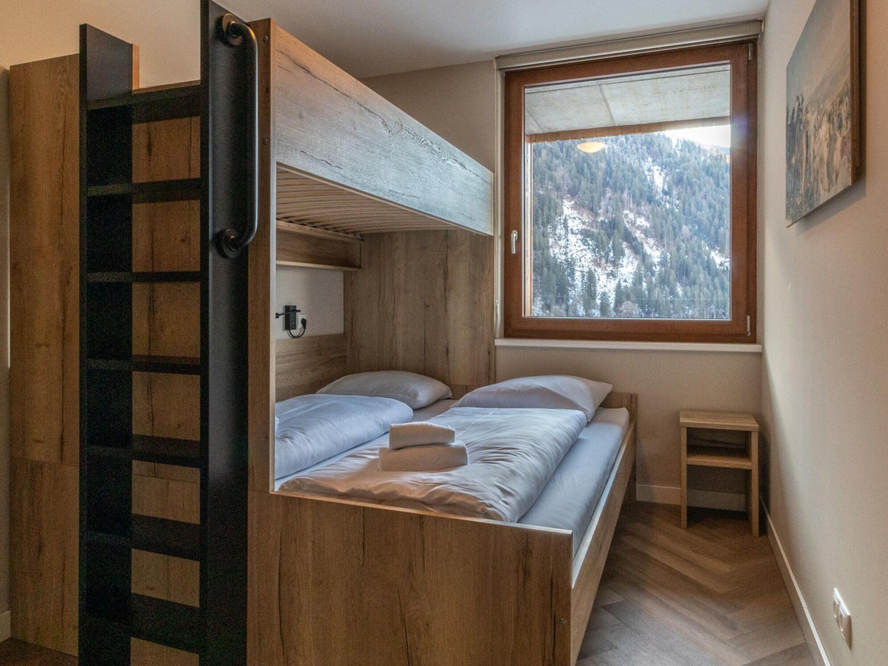 A compact room with a bunk bed, a small window with a view of a forest, and a wooden ladder.