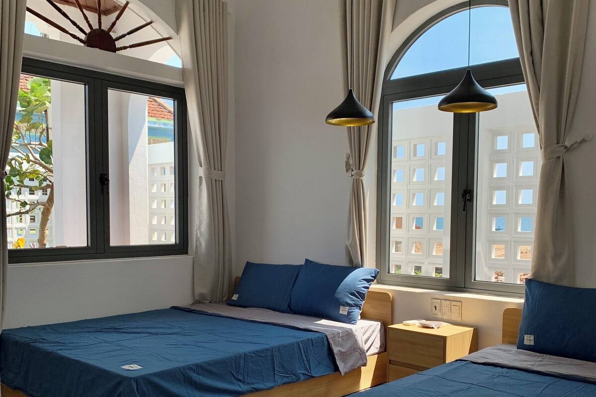 Quarto de hotel com duas camas, uma mesinha e uma janela com cortinas.