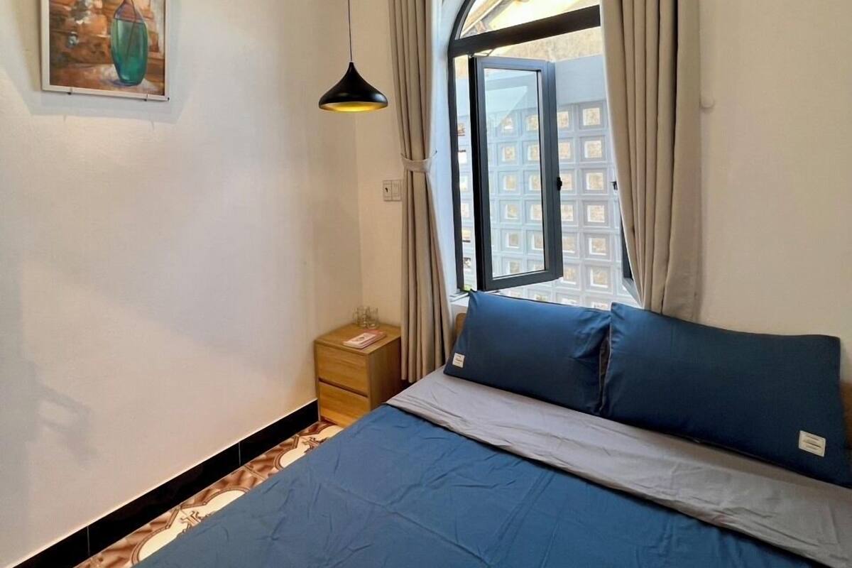 Quarto com cama, mesa de cabeceira, um quadro, janela com cortinas e ar condicionado.
