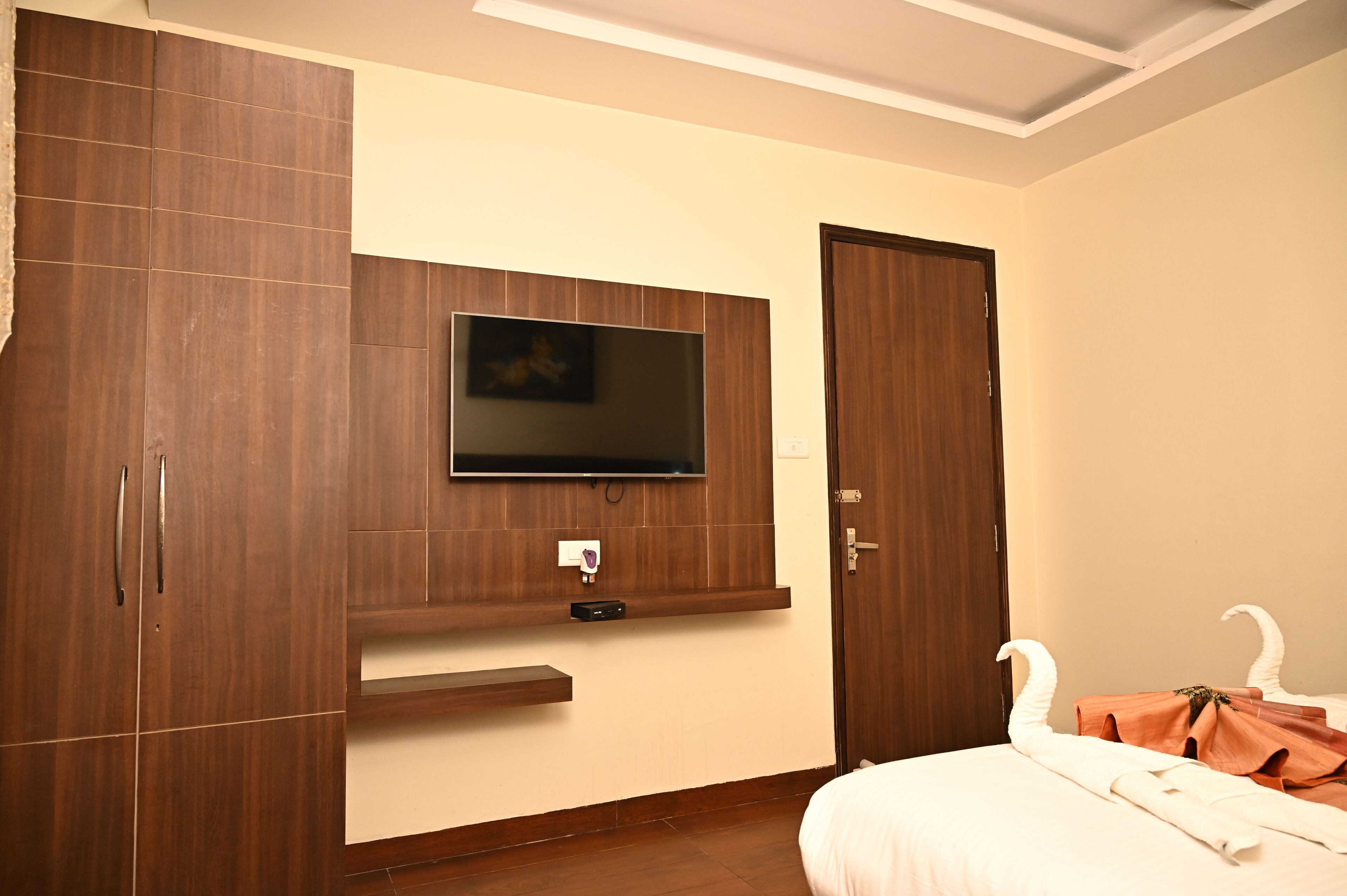 A bedroom with a TV mounted on the wall, wooden wardrobe, and a bed with a white comforter.