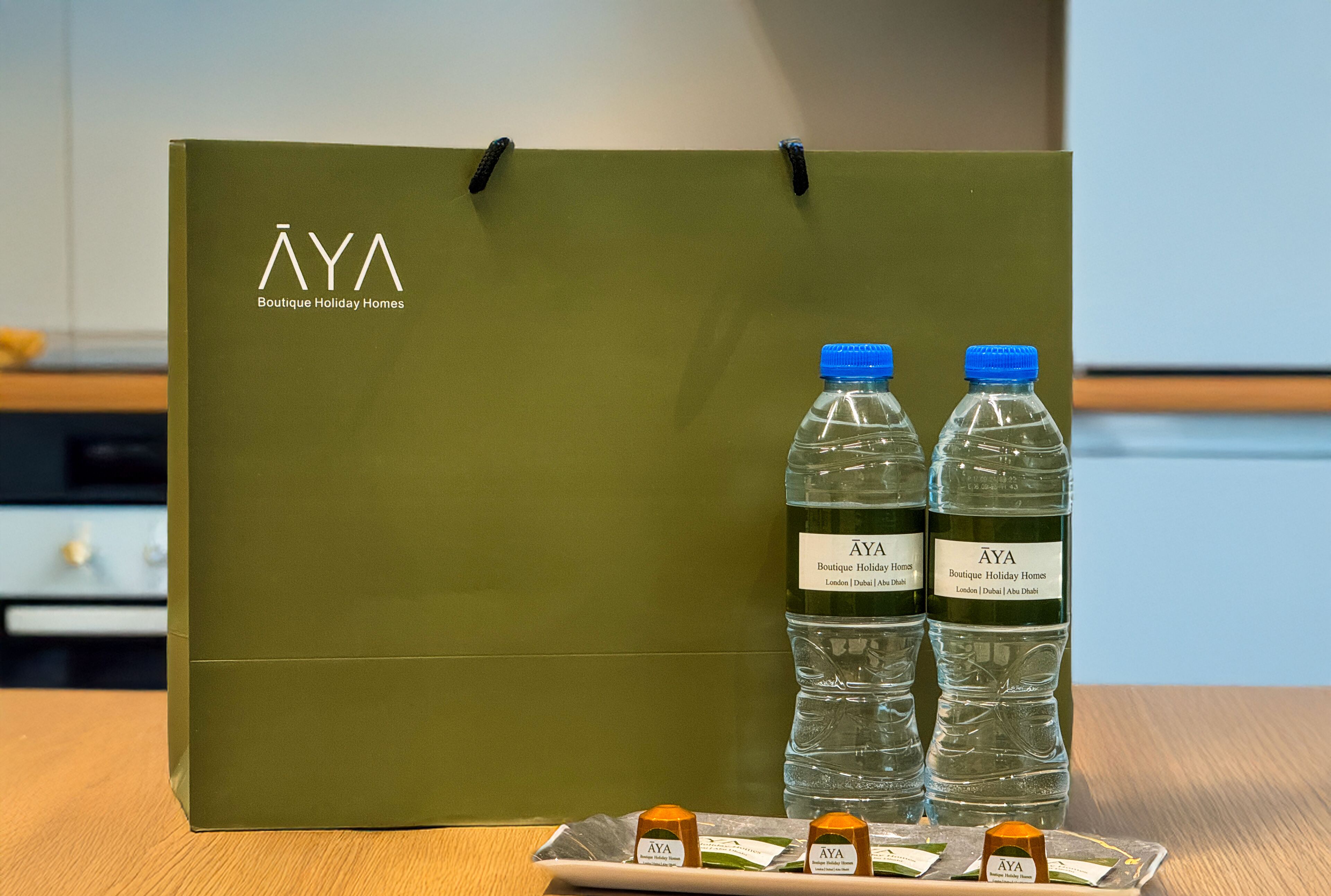 A green AYA Boutique Holiday Homes bag, two water bottles, and three honey jars on a table.