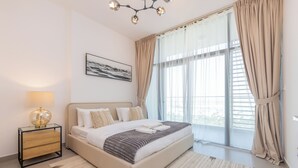 Un dormitorio moderno con una cama grande, una mesita de noche con lámpara, un cuadro enmarcado en la pared y vistas a través de un ventanal con cortinas.