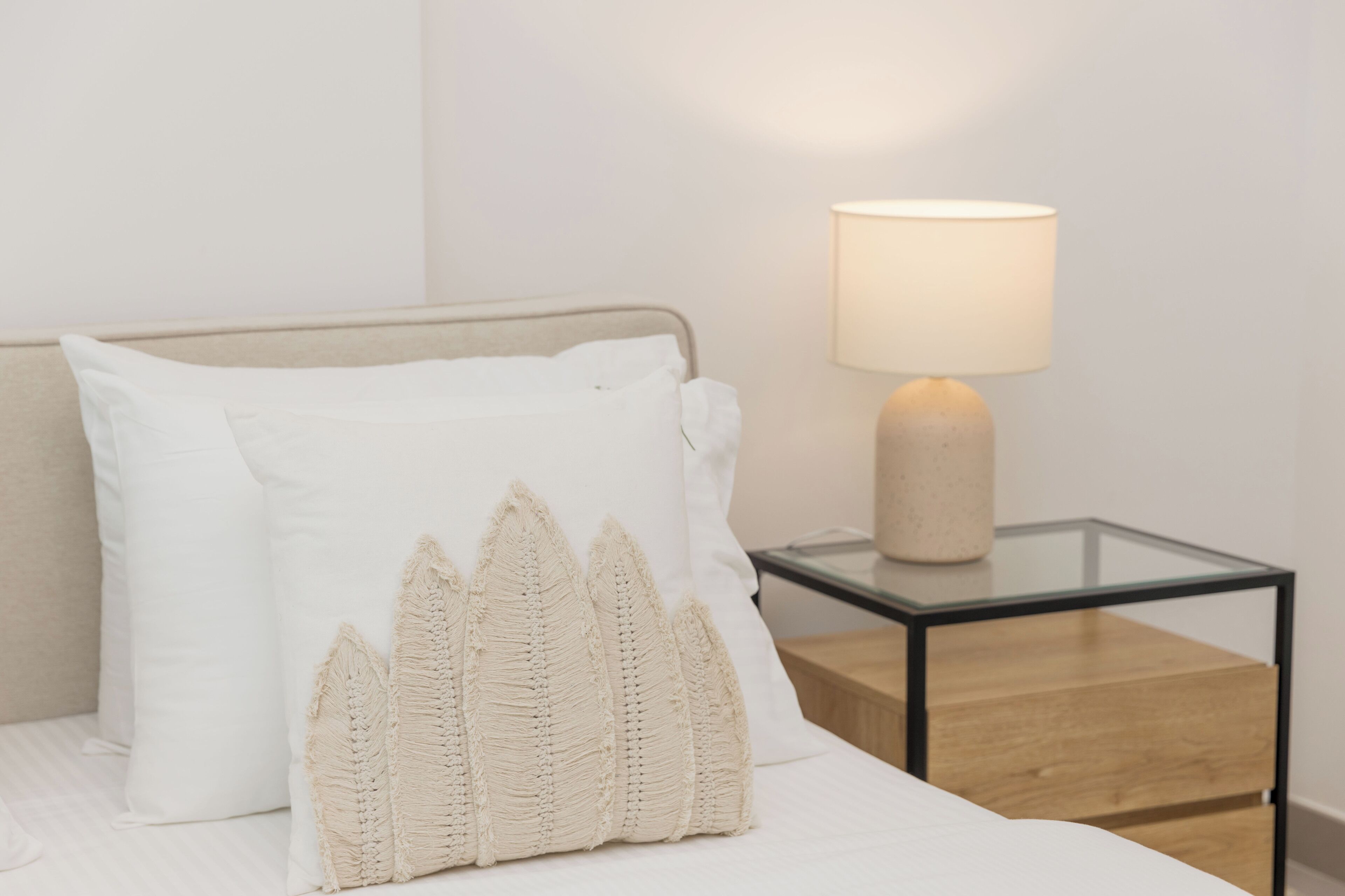 Een net opgemaakt bed met witte lakens en een decoratief kussen. Een nachtkastje met een lamp is zichtbaar.