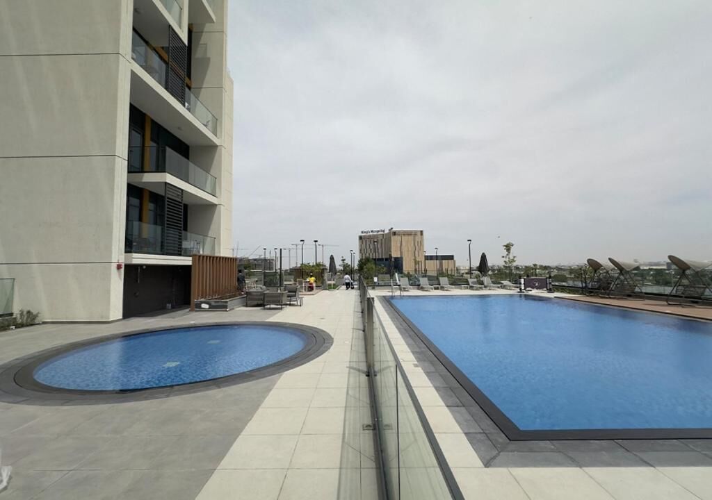A rooftop swimming pool area with two pools, a modern building with balconies, and a distant view of a hospital.