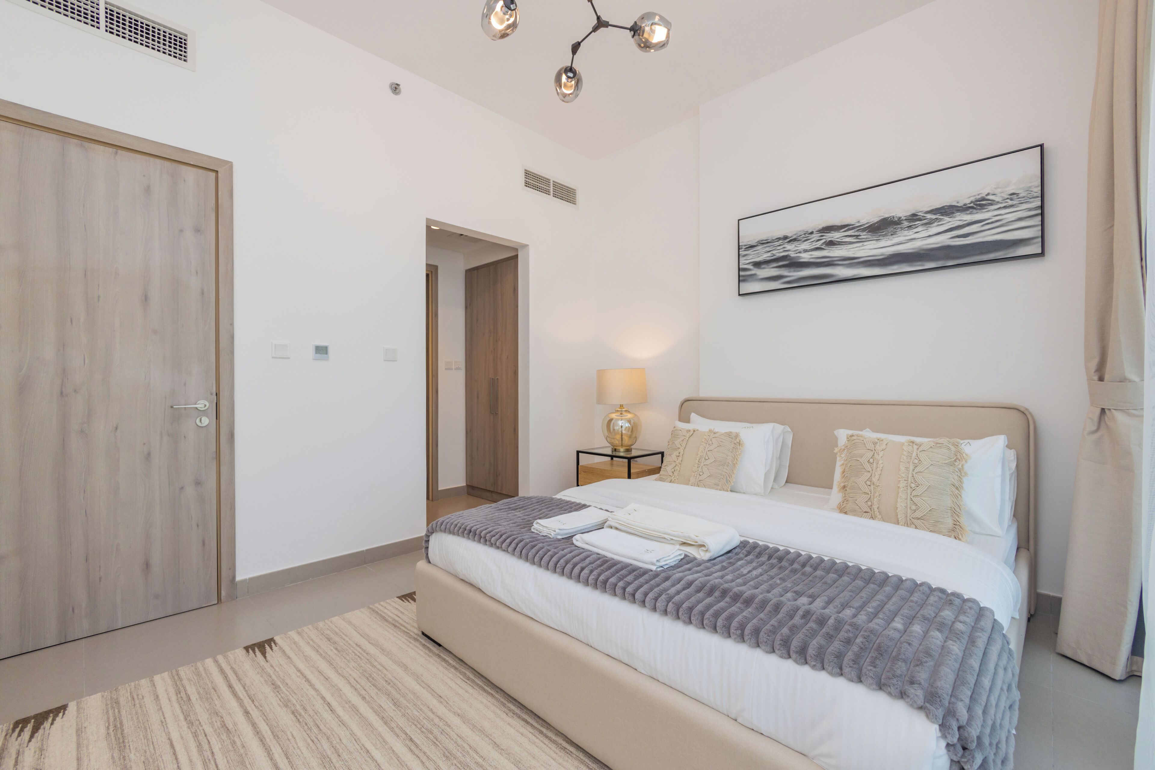 A modern bedroom with a large bed, a bedside table with a lamp, a door, and a painting on the wall.