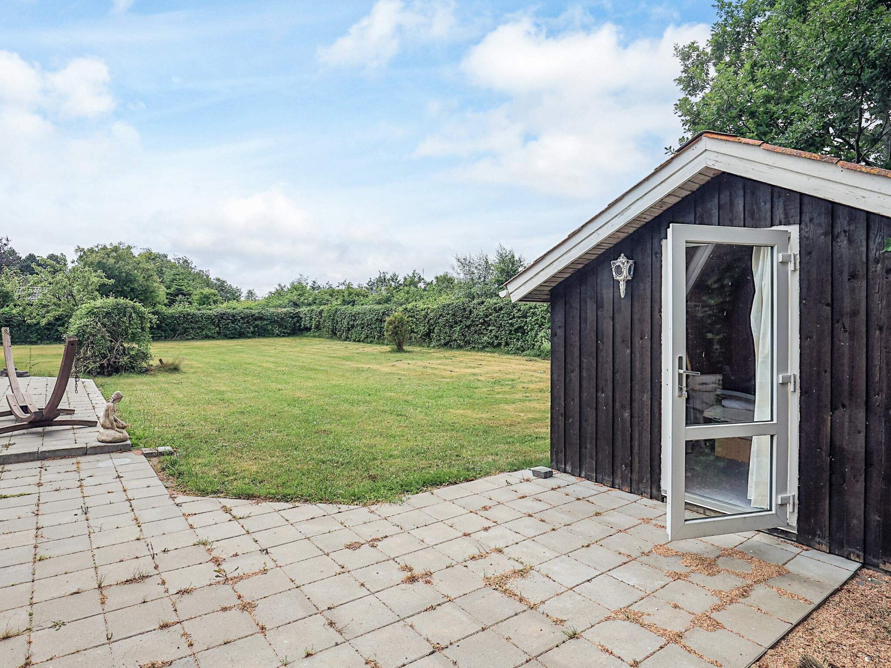 Ein Holzschuppen mit Glastür, eine gepflasterte Terrasse und ein Rasen mit Bäumen im Hintergrund.