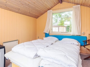 Quarto com teto de madeira, uma cama de casal com roupa de cama branca, janela com cortinas e uma pequena mesa com uma cadeira.