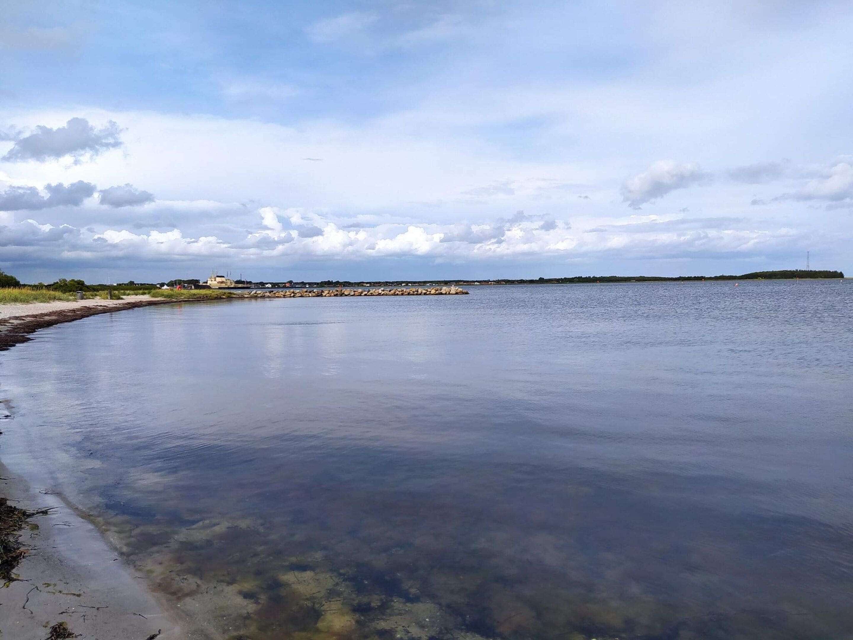 A calm waterfront with a clear sky, a rocky breakwater, and a distant island.