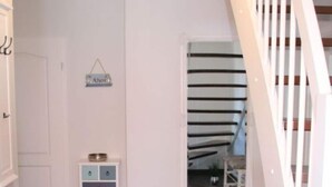 Un pasillo con suelo de madera, una puerta blanca, un pequeño armario azul con cajones y un ventanal con vistas a una escalera.