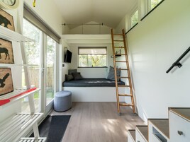 A compact room with a loft bed, a ladder, a small ottoman, and a window with a view of trees.