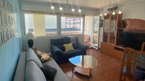 Un salon avec un canapé, une table basse et un téléviseur. De grandes fenêtres offrent une vue sur l’extérieur.