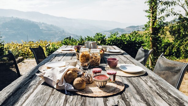 A wooden table set outdoors with various food items and drinks, surrounded by chairs, with a scenic view of mountains in the background. - Quinta De Guimaraes (Baiao)