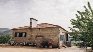 A stone building with a tiled roof, a chimney, and a small staircase leading to a door. There are flower pots with pink flowers and a tree to the right. - Quinta De Guimaraes (Baiao)