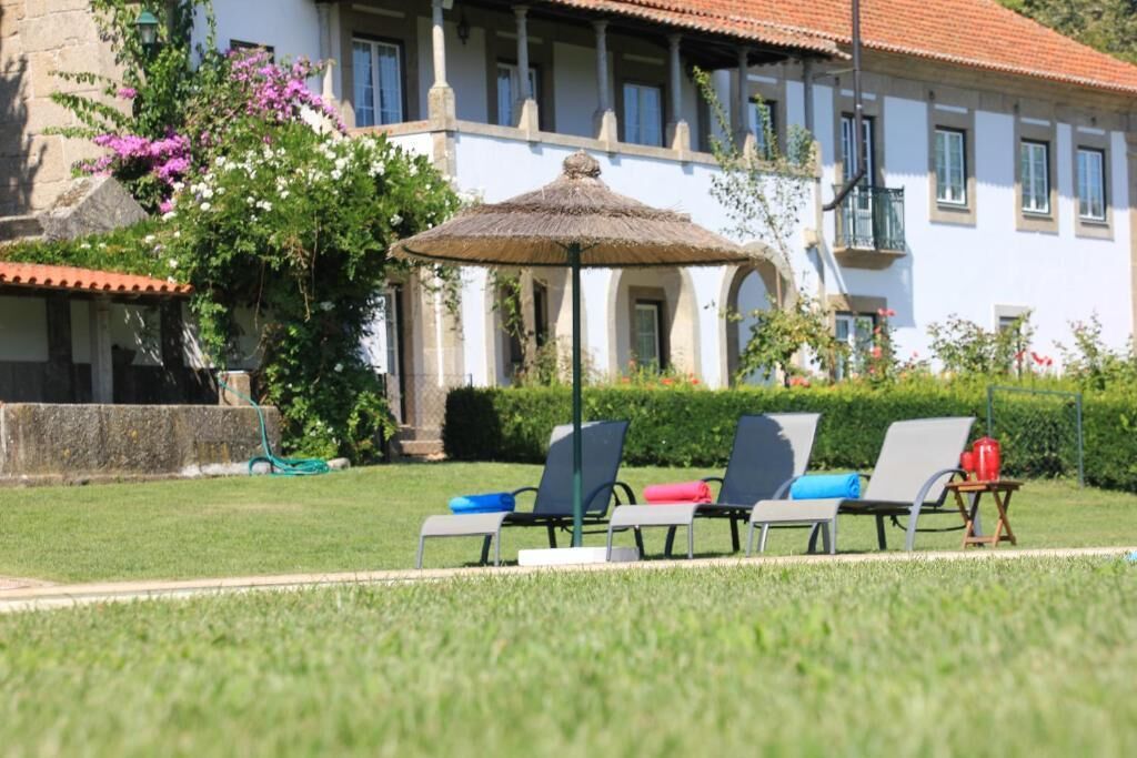 Un edificio con tejado de tejas, un jardín con césped y una sombrilla de paja. Hay sillones con cojines.
