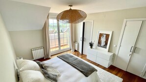 Une chambre avec un grand lit, une commode blanche, un luminaire suspendu et une vue sur l’extérieur par une fenêtre.