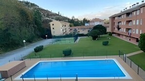 Una piscina amb dues zones separades, envoltada per gespa ben cuidada i una tanca. Al fons, hi ha edificis d'alguns pisos i una carretera.
