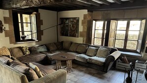 A rustic living room with a large L-shaped sofa, a round coffee table, and a striped armchair. There are multiple windows with wooden shutters, a painting on the wall, and a stone wall. - Spectacular house in Castaneda (Castaneda)