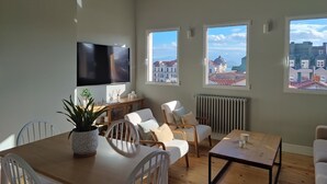 A spacious living area with a large TV, a dining table, and multiple windows offering city views.