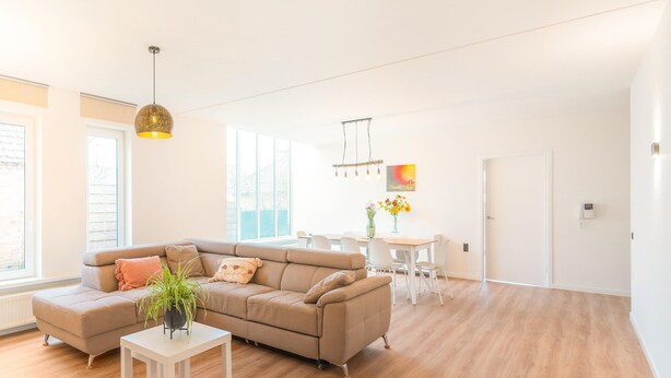 A spacious living area with a beige sectional sofa, a white coffee table, and a dining table with chairs. There is a large window providing natural light and a view of the outside.