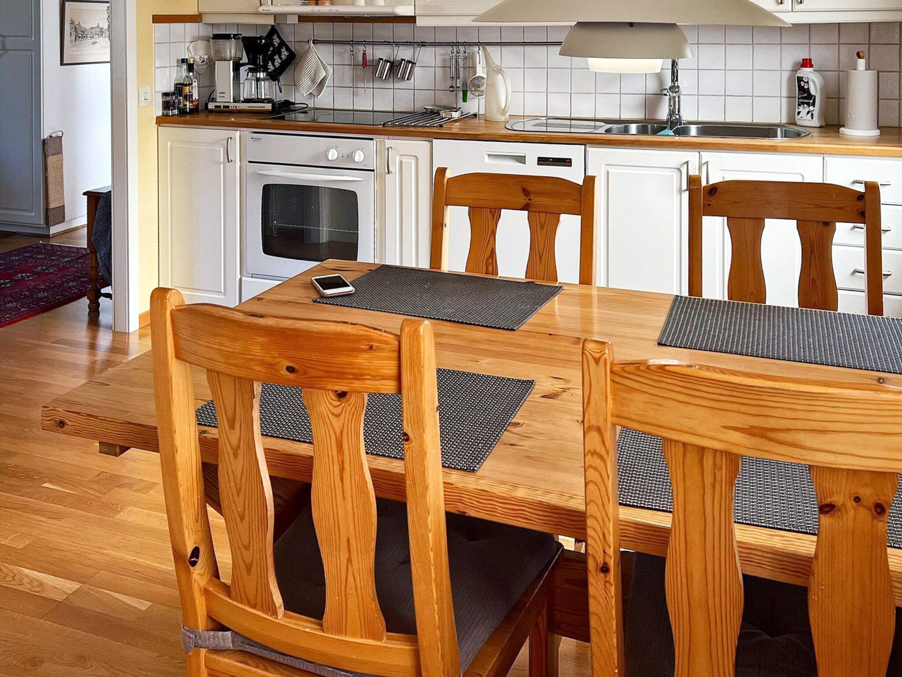 A dining area with a wooden table and chairs, a kitchen in the background, and a red rug on the floor.