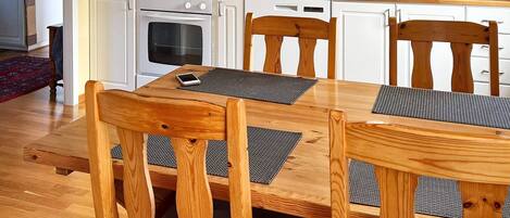 A dining area with a wooden table and chairs, a kitchen in the background, and a red rug on the floor.